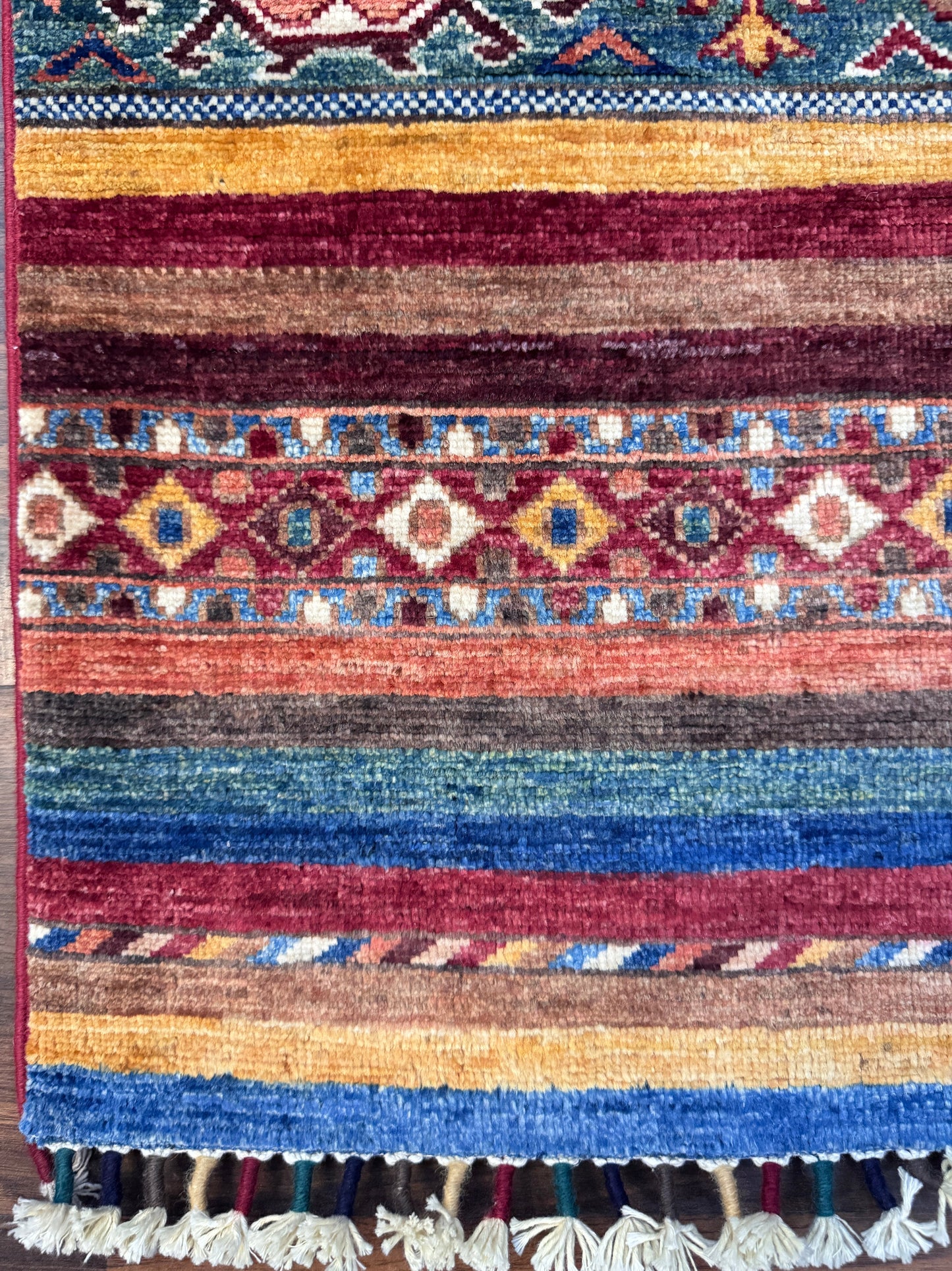 A close-up of a handwoven geometric area rug with horizontal stripes and diamond motifs in blue, red, brown, beige, and yellow, finished with a cream tassel fringe.