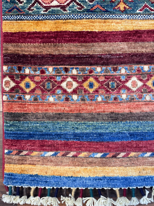 A close-up of a handwoven geometric area rug with horizontal stripes and diamond motifs in blue, red, brown, beige, and yellow, finished with a cream tassel fringe.